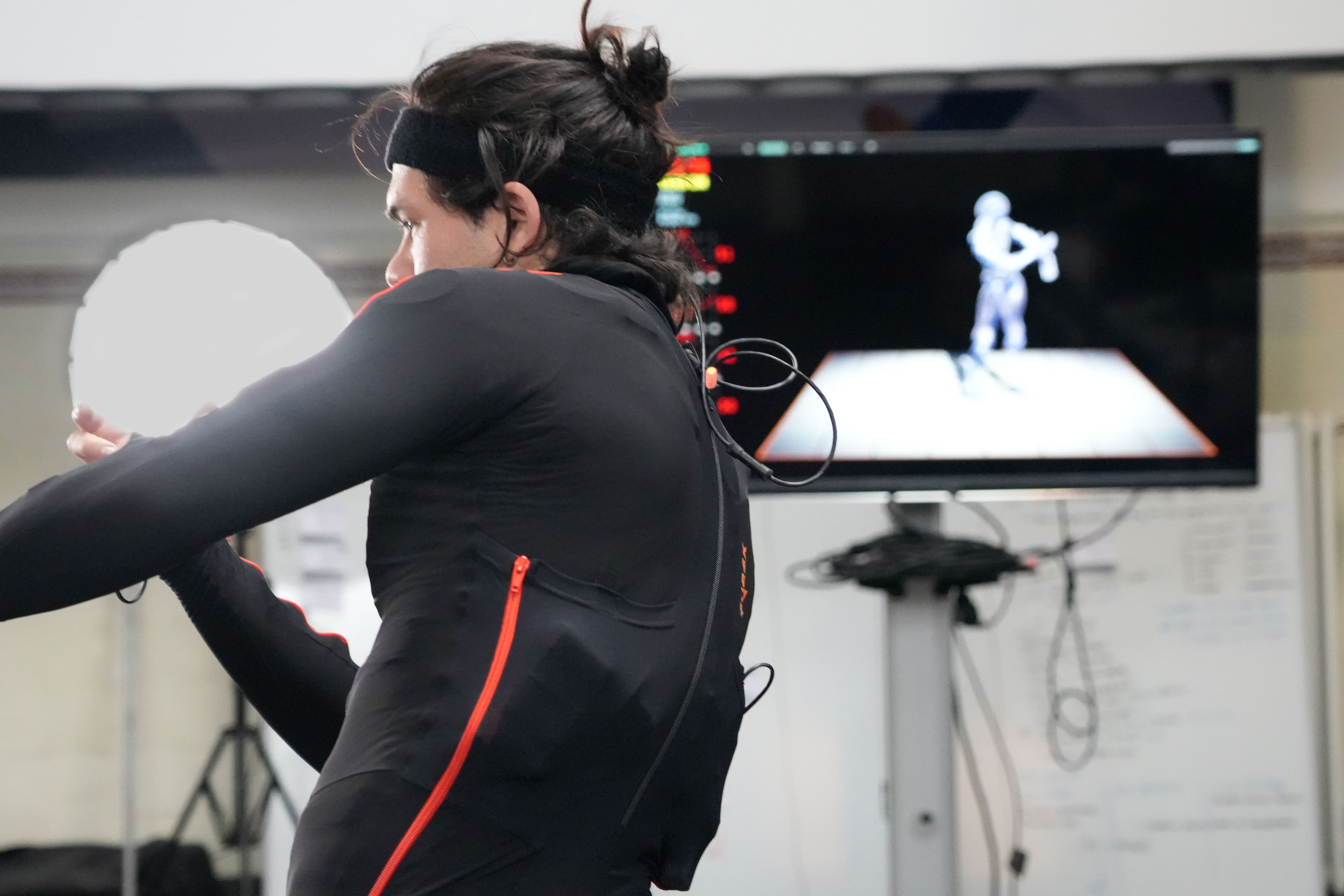 Danser in een motion-capture pak, en een scherm met zijn avatar op de achtergrond.