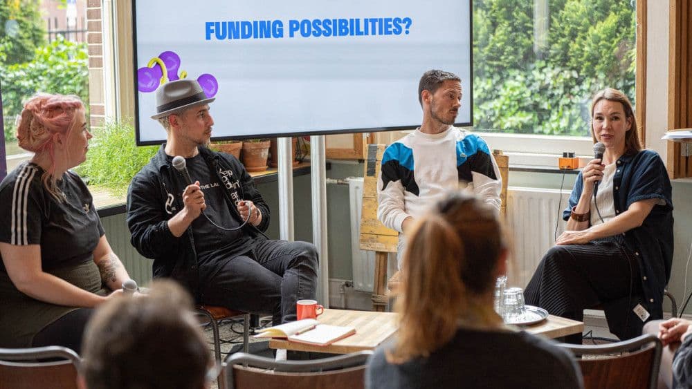 Een panel van vier mensen zit voor een scherm met de tekst 'funding possibilities'. Op de voorgrond silhouetten van publiek. 