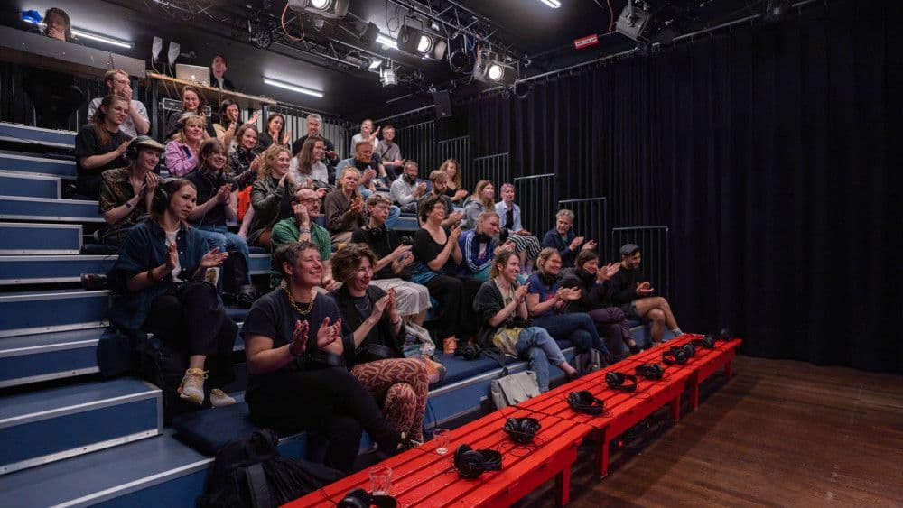 Een tribune vol mensen applaudiseert. Sommigen hebben koptelefoons op, bij anderen ligt die naast of onder hun zitplaats. 